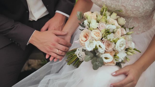 The bride and groom sit and gently stroke their hands to each other. Closeup of hands and wedding bouquet.