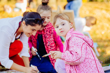 Young attractive mother teaches children painting in autumn park. Creative child painting on nature sitting on ground. Outdoors activity for school age children concept