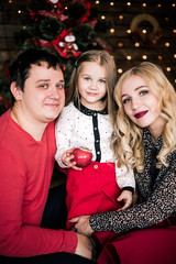 Beautiful young family in red having fun together for Christmas holidays, sitting on a living room floor next to a nicely decorated Christmas tree