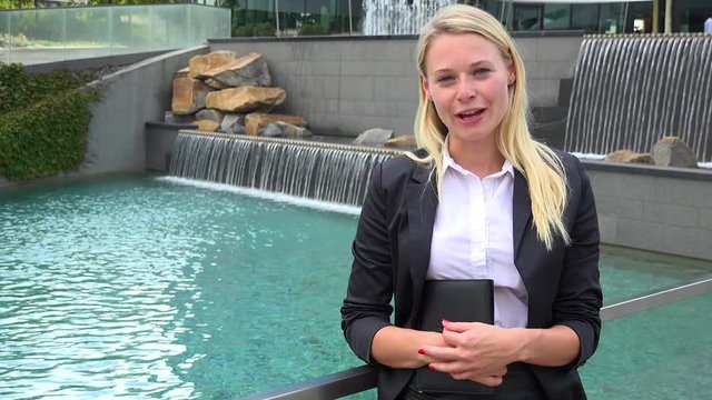 A young beautiful businesswoman talks to the camera in an urban area - artificial waterfalls and a pool in the background