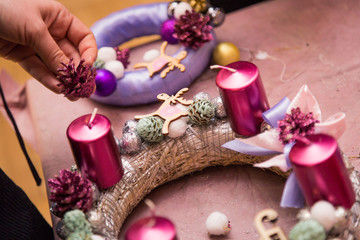 Christmas decoration round wreath, shiny accessories, candles and cones, golden, balls, on pink wrapping paper, creating process with hands