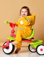 Baby girl kid riding her first bicycle tricycle in warm yellow overalls looking up on light grey 