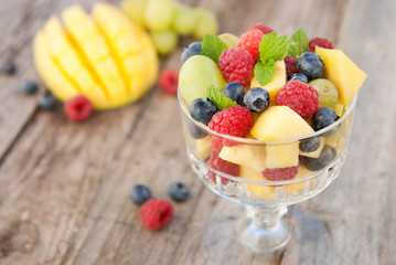 Delicious fruits salad in glass - raspberry, mango, blueberry, grapes, mint leaves. Wooden table, rustic background.