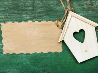 Small wooden bird house with heart decoration and blank paper on green retro wooden background,...