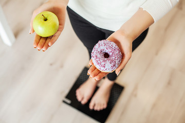 Diet. Woman Measuring Body Weight On Weighing Scale Holding Donut and apple. Sweets Are Unhealthy...