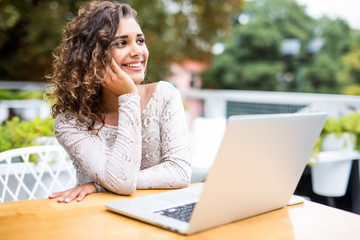 Portrait of curly latin woman in glasses working with her laptop outdoors. Charming freelancer woman with curly hair is sitting in street bar and doing her remote work on the netbook