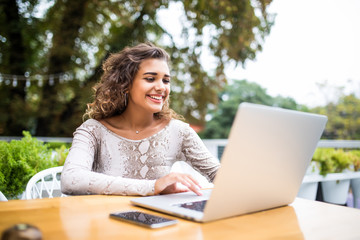 Portrait of latin beautiful woman in glasses working with her laptop outdoors. Charming freelancer woman with curly hair is sitting in street bar and doing her remote work on the netbook