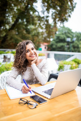 Smiling latin female student doing homework by laptop at cafeteria table