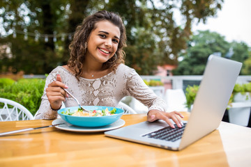 Curly latin woman hold cutlery eat salad in cafe restaurant lunch a laptop on the table in a cafe