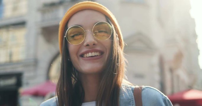 Close up of the attractive Caucasian cheerful girl in sunglasses and a hat looking at the side and then laughing to the camera. Outdoors. Portrait.