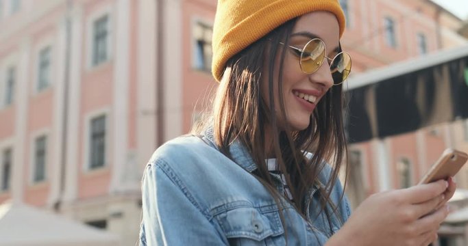 Close up of the Caucasian stylish girl in sunglasses and a hat scrolling and tapping on the smartphone outdoor. Portrait.