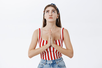 Faithful sexy adult woman in striped top and headband holding palms in pray against chest raising head up looking in sky praying being concerned asking god help, posing against grey wall