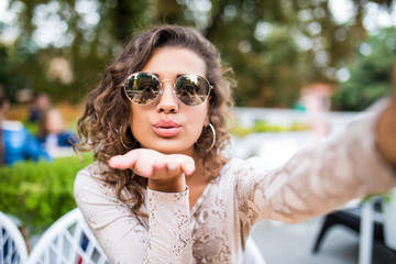 Attractive young latin woman in sunglasses with curly hair takes a selfie, posing and looking at camera, outdoors.