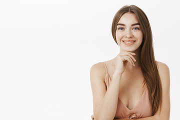 Waist-up shot of sensual and feminine brunette in cute beige dress holding hand on chin and smiling broadly having pure clear skin delighted to have no problems using minimal makeup over grey wall