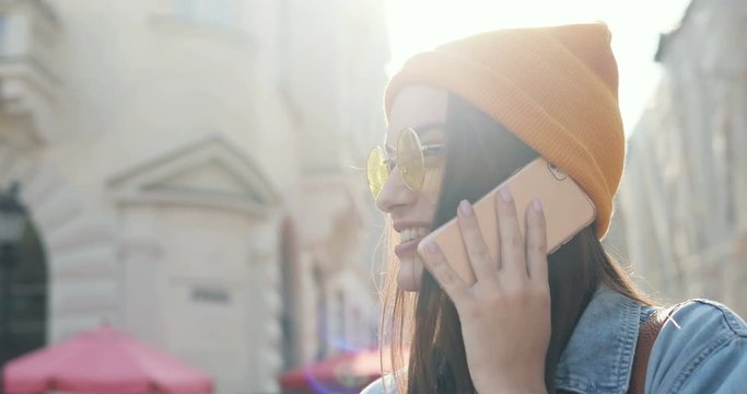 Young stylish beautiful Caucasian woman in sunglasses anda hat talking on the mobile phone cheerfully in sunlight on the street. Outdoors.