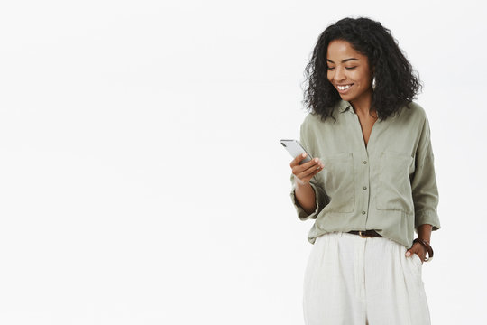 Stylish and successful dark-skinned female african entrepreneur checking mail box on way to office holding hand in pocket standing casually over gray wall holding smartphone smiling at gadget screen