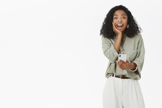 Girlfriend Receive First Payment On Online Bank Account Rejoicing Feeling Happy And Surprised Touching Cheek Looking With Joy And Amazement At Camera Holding Smartphone Posing Against Gray Background