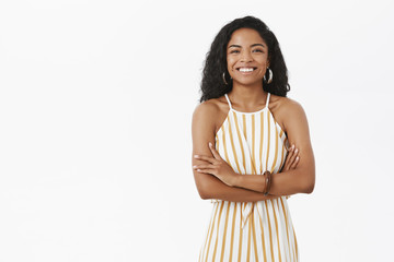 Indoor shot of elegant successful charming african american businesswoman with curly hairstyle in yellow striped dungarees crossing arms on chest in self-assured gesture smiling with delight