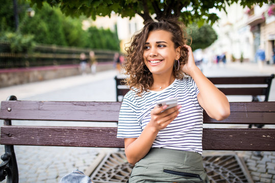 Young beauty curly latin woman typing on the phone while sitting on the bench on the street