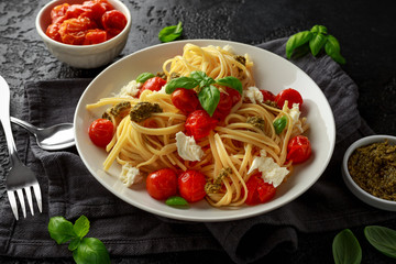 Pasta with green pesto sauce, roasted cherry tomatoes and mozzarella cheese in white plate on dark rustic background
