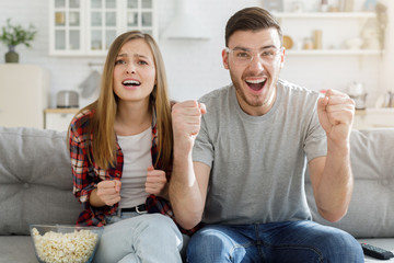 Young couple watching tv game, sitting on sofa at home, cheering for team, supporting together