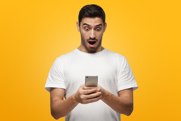 Young man looking at display of his phone with mouth open, isolated on yellow background