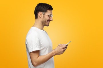 Young attractive man looking and at smartphone while texting, using mobile phone, isolated on yellow background