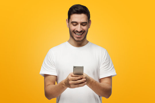 Indoor picture of handsome Caucasian man isolated on gray background wearing white casual T-shirt laughing happily being amused by content on screen of smartphone he is holding in both hands