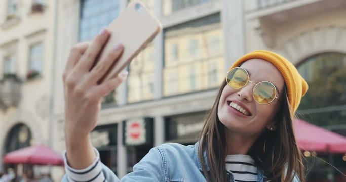 Close up of the emotional cheerful Caucasian stylish girl in sunglasses and hat taking cool selfies on the smartphone camera on the street. Outdoor. Portrait.