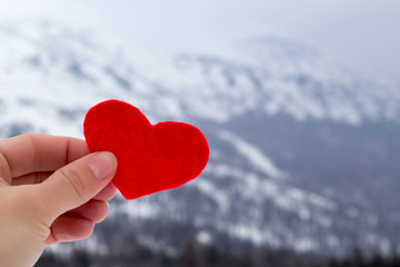 Fototapeta premium Hand holding red felt heart on the winter mountains landscape snow covered background. Love winter, ski concept. Copy space