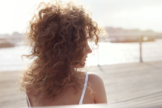 Authentic vivid profile portrait of young Caucasian beautiful female, admiring sunset on city sky, looking afar. Wind is messing long curly hair. Close up shot of woman with river side on background.