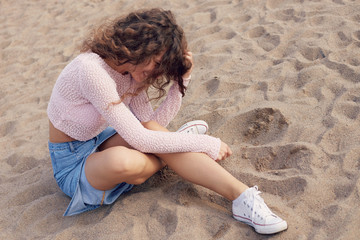 Young cheerful curly haired lady chilling on beach, laughing , flirting with lover, having carefree stylish casual look, dressed in denim skirt, pink top and white sneakers. Outdoor activities concept