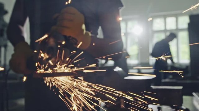 Man cutting metal with angle grinder, working indoors in workshop. Bright sparks are visible in close-up.