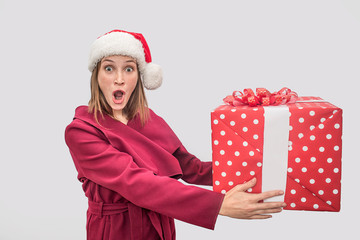 Wondered and amazed young woman in red coat and Christmas hat holding big box of present. Model...