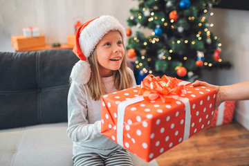 Obraz na płótnie Canvas Beautiful little girl holds present with both hands and looks up. She is excited and happy. Adult holds present as well. There are Christmas tree and sofa on background.