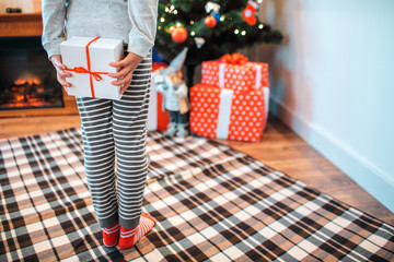 Cut view of small girl standing back to camera. She hides box of white present. Girl stands in front of Christmas tree with gifts.