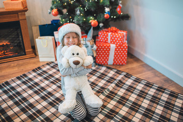 Happy and positive girl sitting on blanket on floor and embacing white toy. She smiles. Girl looks happy. She in room alone.