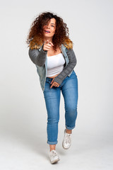 Body positive concept. A smiling, curly plus size girl, dressed in a casual style, points her finger at the viewer. White background, studio.
