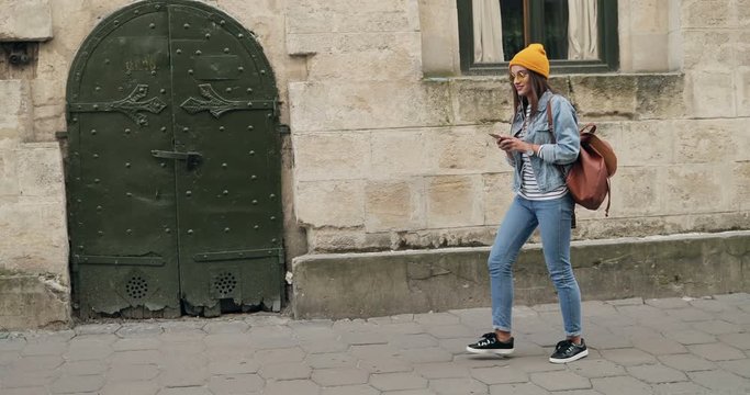 Young Caucasian stylish woman in the glasses, hat and with a backpack walking the town street and listening to the music on the smartphone device via headphones. Outdoors.