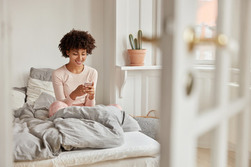 Positive dark skinned female reads internet publication on website via cell phone, watches video, connected to wireless internet, poses in nightwear on comfortable bed, has spare time and weekend - obrazy, fototapety, plakaty