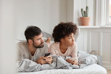 Contemplative family couple use cellulars, concentrated aside, lie in bed at morning, connected to wireless internet at home, text messages, invite friends for party, ignore live communication