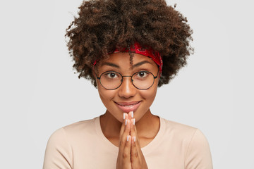 Photo of pleasant looking dark skinned girl keeps hands together near mouth, looks directly at...