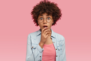 Fototapeta na wymiar Isolated shot of pretty black woman has curly hairstyle, horrified to find out friend is in hospital, keeps hand near opened mouth, wears round glasses and denim jacket, isolated over pink background
