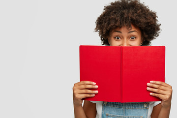 Indoor shot of of cheerful woman covers face with red textbook, has joyful expression, being in...
