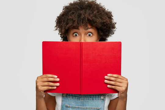 Shocked student with Afro haircut, holds opened red book in front, covers half of face, has deadline to prepare for exam, keeps eyes widely opened, models against white studio wall, reads literature