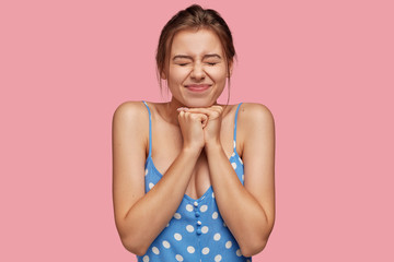 Hopeful dreamy woman keeps eyes closed, hands pressed together, anticipates something wonderful, dressed in blue polka dot clothes, models against pink background. Student waits exam results