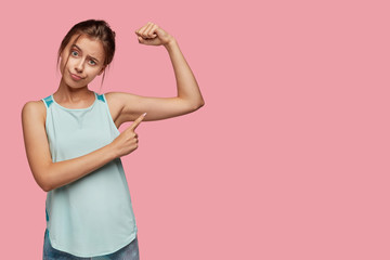 Healthy lifestyle and sport concept. Dissatisfied European woman points at arm muscle, wants to have muscular body, poses over pink background with free space for your advertisement or promotion