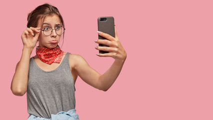 Upset gloomy girlfriend purses lips, makes selfie portrait with modern cell phone, keeps hand on...