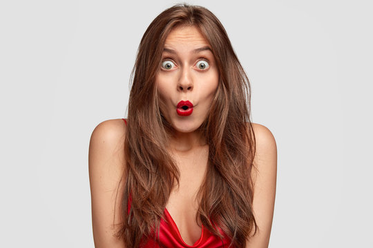Image of shocked green eyed young female keeps mouth round, has red lipstick, long straight hair, stupefied expression, poses over white background. People, reaction and facial expression concept
