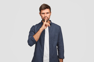 Photo of serious male keeps index finger on lips as demonstrates hush gesture, asks be quiet, dressed in fashionable clothes, angry as someone tells his secret, isolated over white background